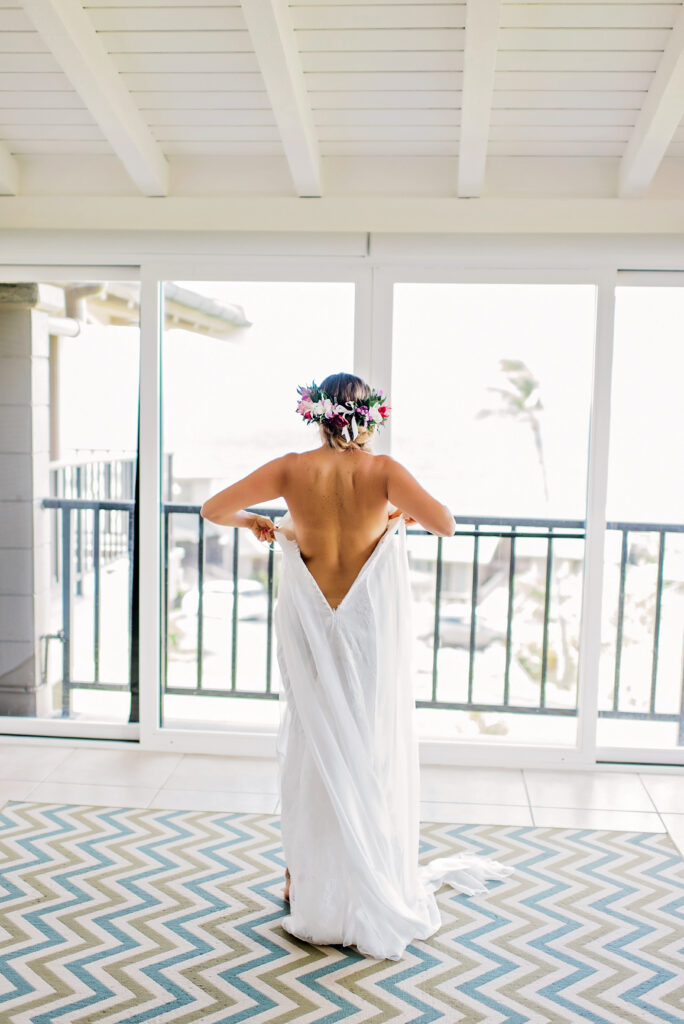 Bride in a simple white gown with a lei, standing on a balcony with her back turned, embracing the moment.