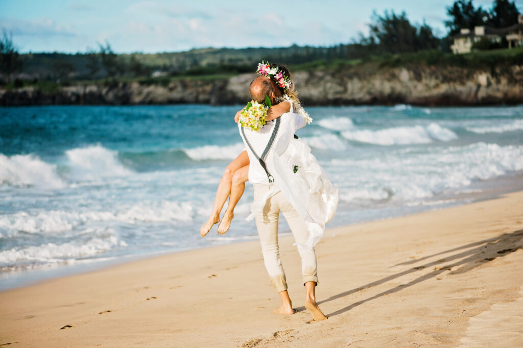 The groom carries the bride laughing as waves lap their feet, embodying carefree elopement.