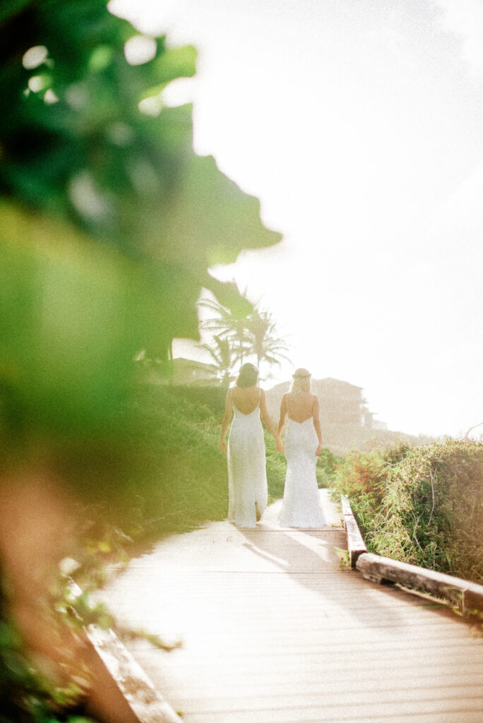Dori & Natalie walk hand in hand down a sunlit wooden pathway, ready to say “I do.”