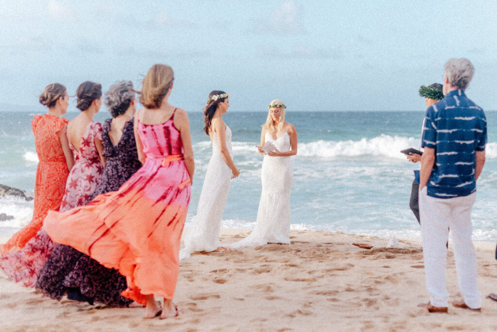 Guests in vibrant dresses watch as the brides say "I do" on Ironwood Beach, Maui.