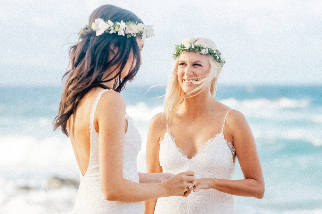 The brides exchange heartfelt vows, ocean waves crashing behind them on their wedding day.