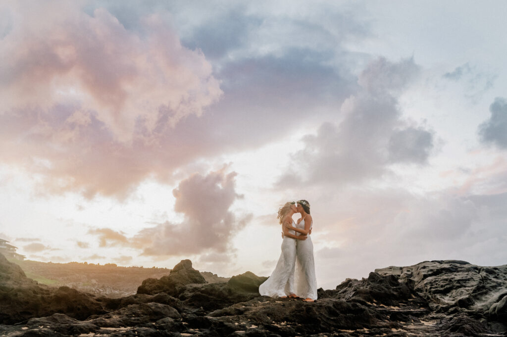 A cinematic moment as one bride stands alone on the rocks, framed by a pastel Maui sky.