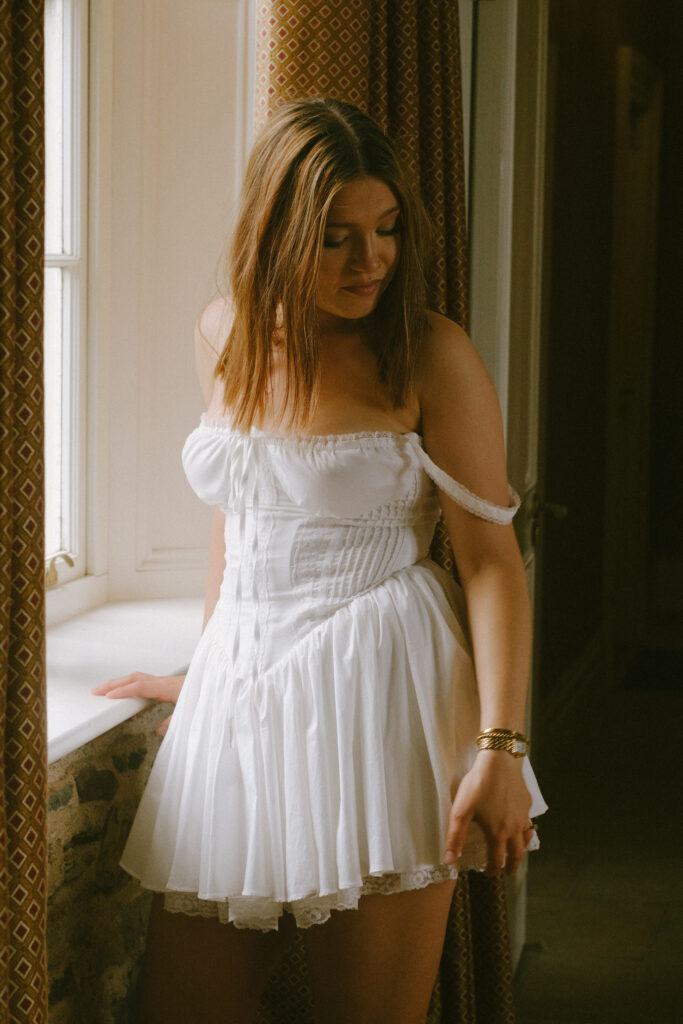 Bride in a white satin robe, standing by a window, illuminated by natural light in Wilton Castle.