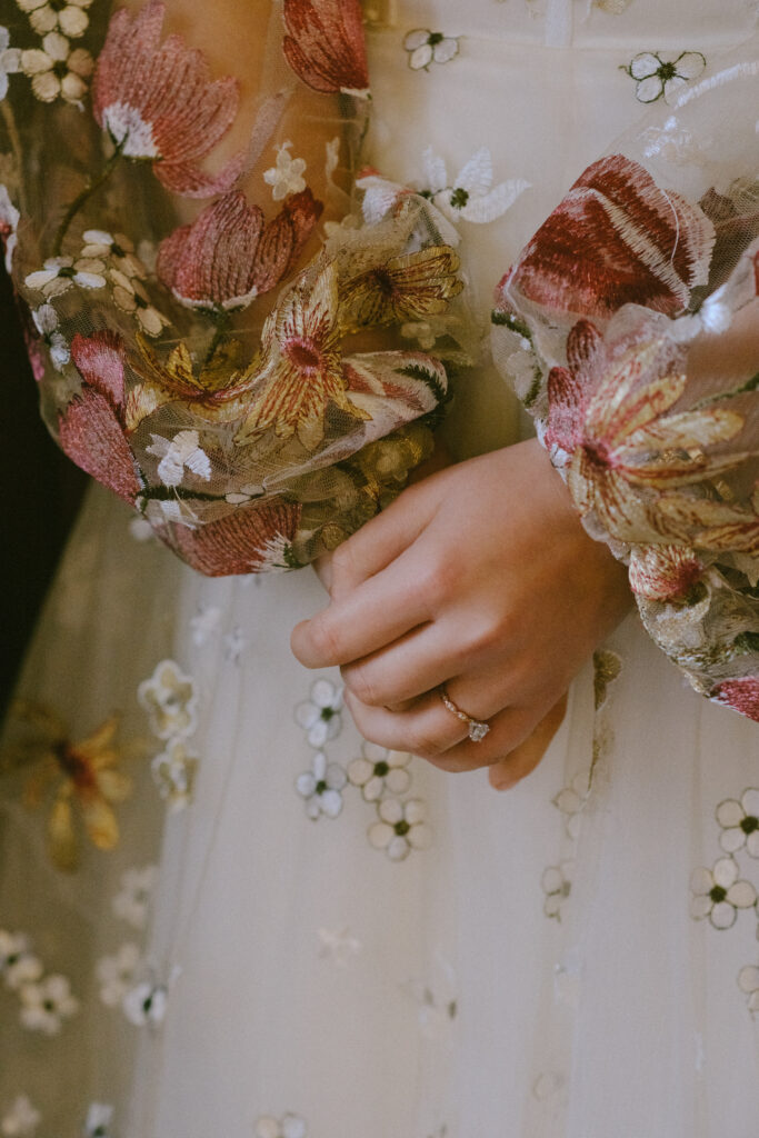 Close-up of bride’s hands showcasing intricate floral dress details and romantic bouquet.