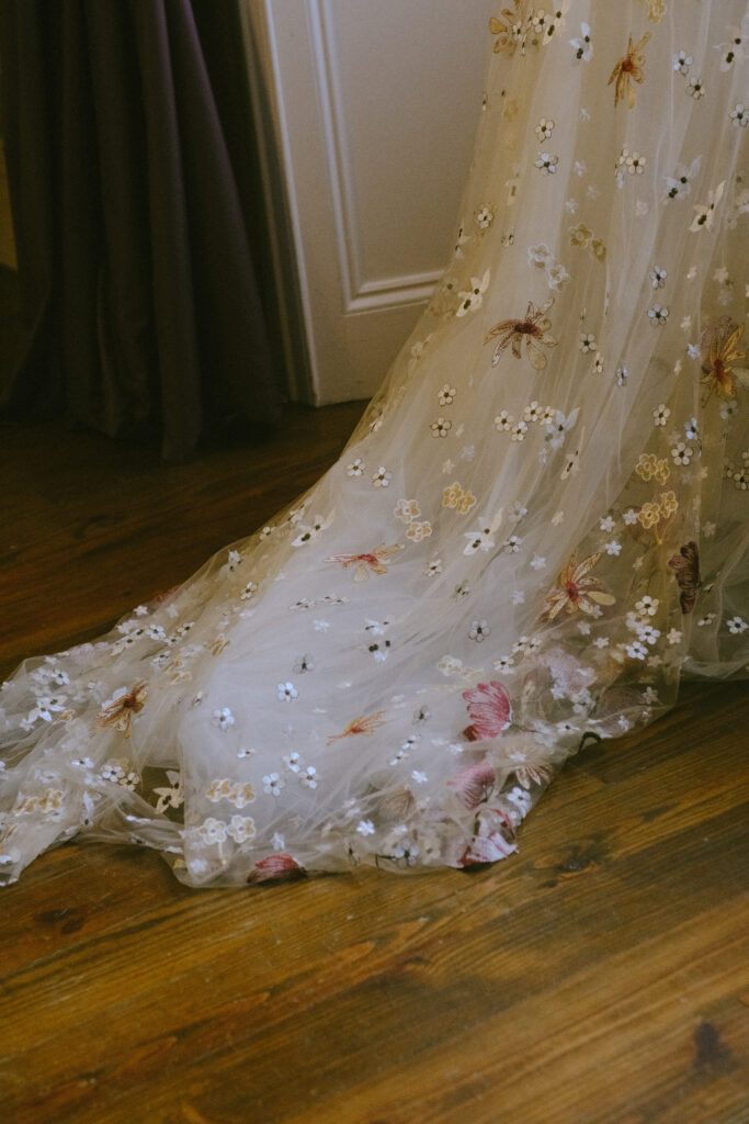 Bride’s train with floral appliqué drapes across a wooden floor, adding elegance to the moment.