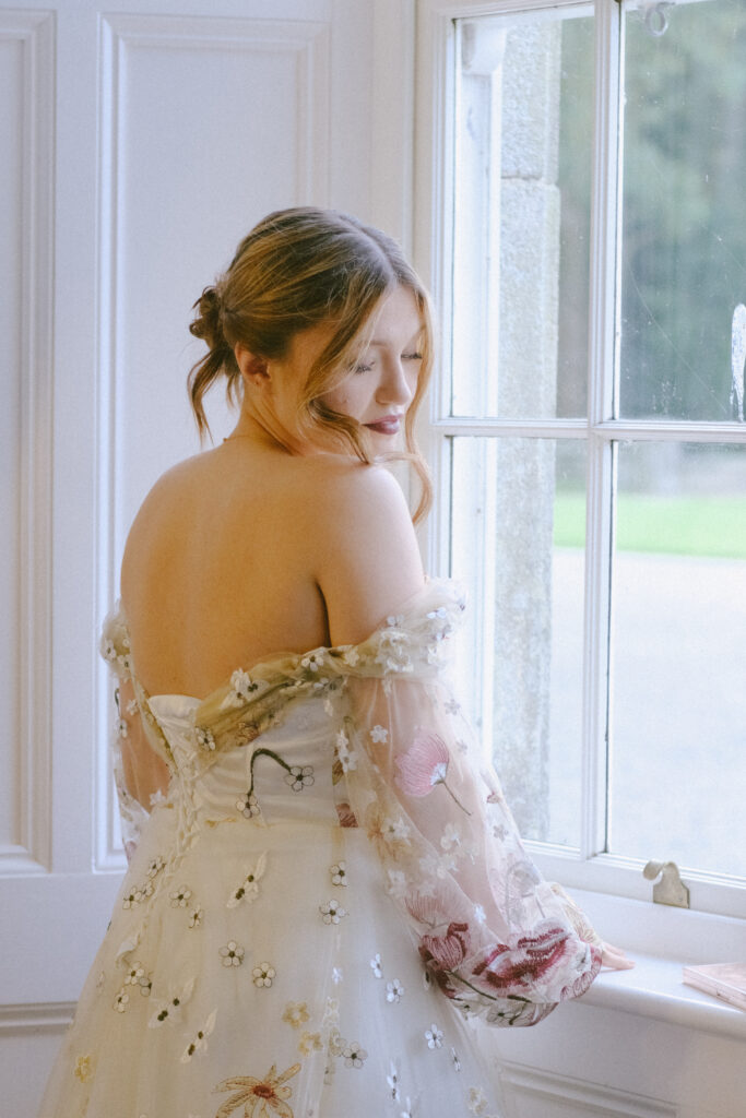 Bride gazes out the window holding her bouquet, bathed in soft natural light at Wilton Castle.