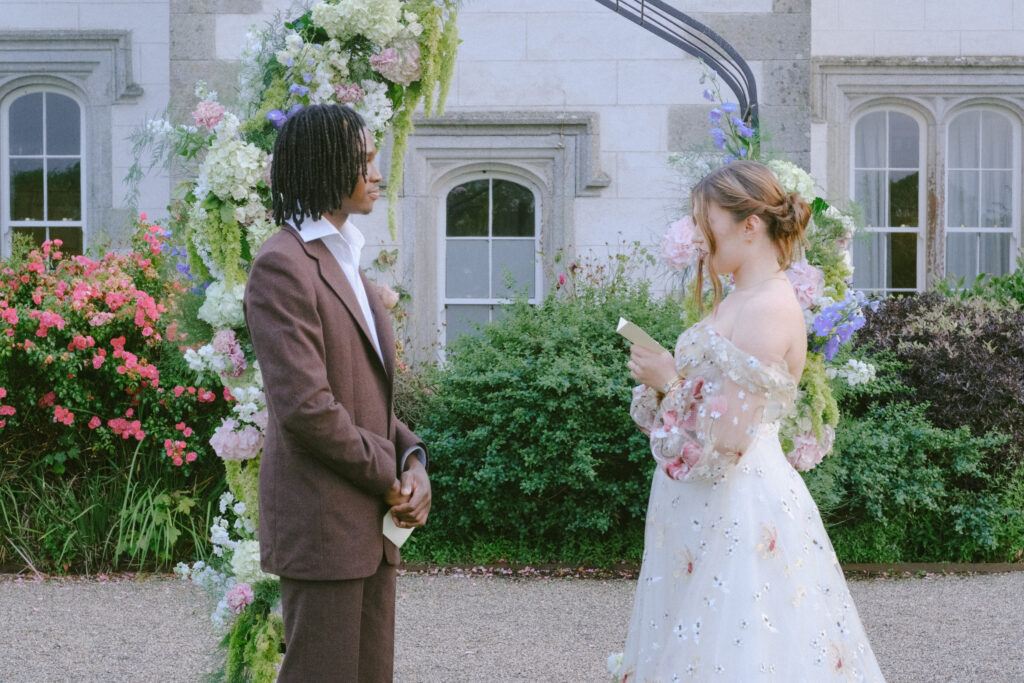 Bride smiles while reading her vows, with the groom lovingly looking at her under a floral arch.