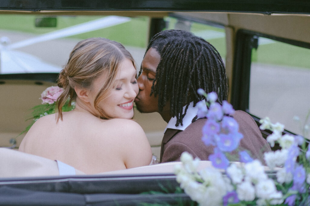 Groom kisses bride while seated in a classic car adorned with fresh florals and greenery.